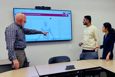 A male professor points to data on a large monitor while students look on.